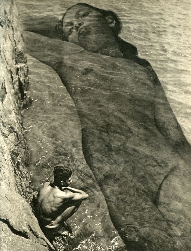 Photogravure. We look down at a calm sea lapping at steep cliffs. A boy squats on what appears to be a natural stone ledge by the water’s edge and peers into the water. At a much larger scale, a young woman’s nude, prone body is overlaid in the water so it spans the height of the picture.