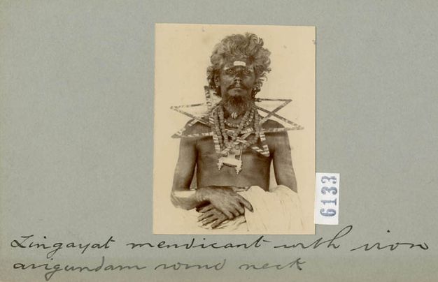 Black and white photograph. Shown from the waist up, a dark-skinned man sits looking at us with his hands clasped in his lap. Flat triangular or star-shaped forms lie around his neck and across his shoulders like stiff, oversized necklaces. The photograph is mounted on a card with handwritten text beneath.