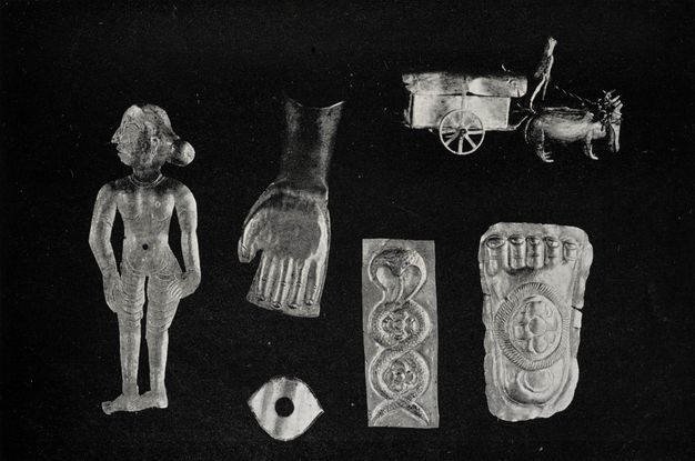 Black and white photograph. Six metal charms are shown against a black background: a woman, a hand, an eye, a cobra, a foot, and a carriage pulled by animals.