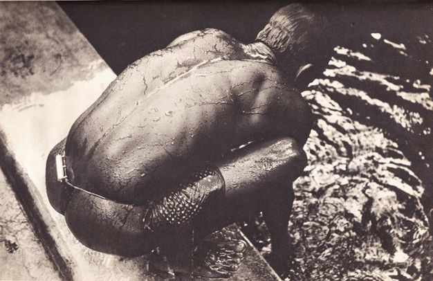 Black and white photograph. The crouching back of a dark-skinned man nearly fills this composition. Water glistens off his skin and runs in a rivulet down the indentation of his spine. He bends forward and reaches into the water beyond the ledge on which he squats.