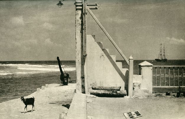 Photogravure. A flight of stairs appears to lead to the top of a telegraph station pole at the centre of this surreal, black and white landscape. A body of water opens beyond the stairs, and on the horizon is a two-masted sailing ship. A goat strolls along a walkway to our left. The cover of a magazine on the ground near us in the lower right corner reads, \“Shaw on Stalin\”.