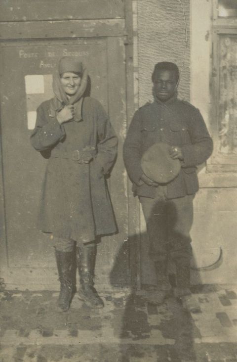 Black and white photograph. A pale-skinned woman and dark-skinned man stand against the side of a building. The woman smiles and holds one hand to the scarf draped over her hat and shoulders. The man holds a round object, perhaps a cap, in his hands. The shadow of the photographer falls over the bottom half of the man’s body.