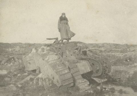 Black and white photograph. A woman stands atop an overturned tank in a rocky landscape. She wears a hat and long coat, which lifts in the wind.
