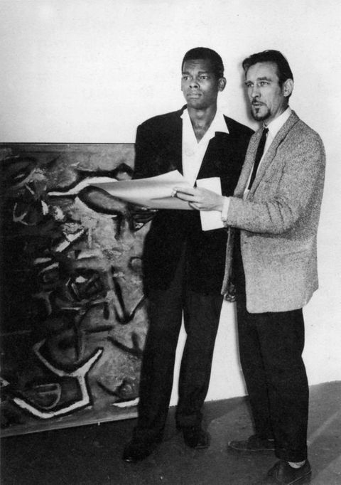 Black and white photograph. Two men stand next to each other. Both wear suits and together hold a piece of paper as they look off to our left. The man to our left, Aubrey Williams, has dark skin and short black hair. The other man, Denis Bowen, has pale skin and a dark goatee.