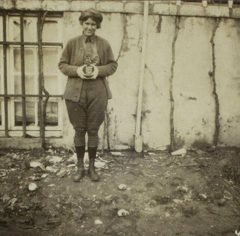 Black and white photograph. A woman smiles at us as she holds a camera in front of her torso. She wears a jacket, button-up shirt, and jodhpur-like trousers over ankle boots.