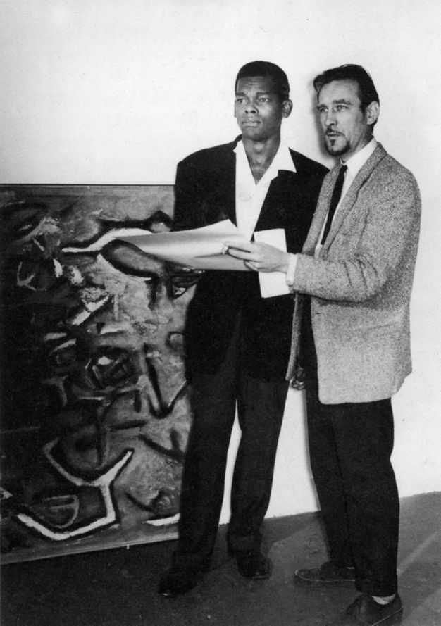 Black and white photograph. Two men stand next to each other. Both wear suits and together hold a piece of paper as they look off to our left. The man to our left, Aubrey Williams, has dark skin and short black hair. The other man, Denis Bowen, has pale skin and a dark goatee.