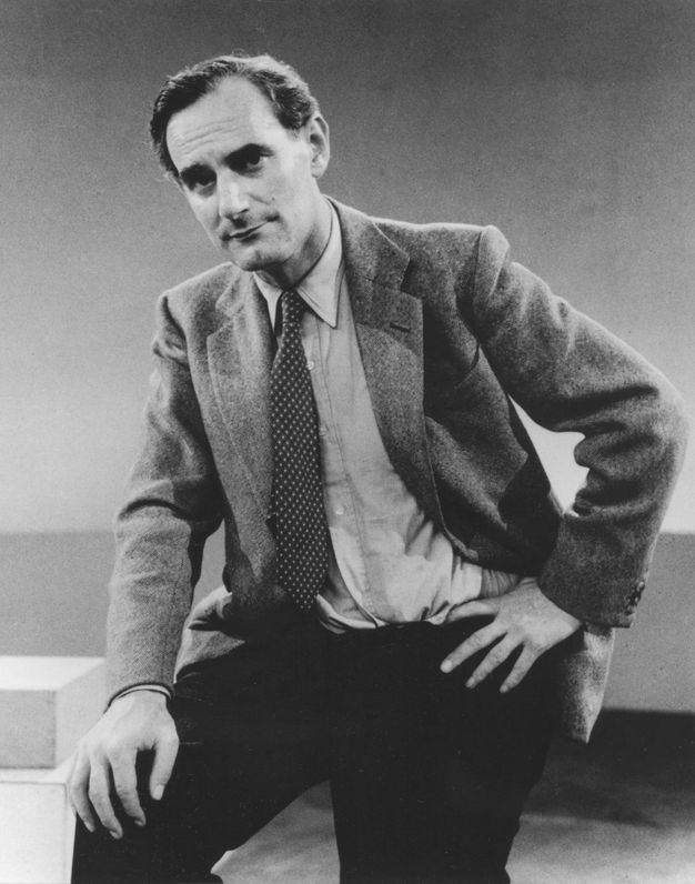 Black and white photograph. Shown from the knees up, a pale-skinned man with dark hair looks at us from under dark brows. He has a square jaw and cleft chin. He wears a suit and holds his jacket open with one hand resting on his hip.