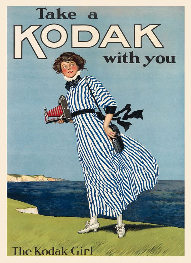 Ad. A pale-skinned woman wearing a long dress with a high, ruffled neck stands near a cliff edge overlooking the sea, a telescoping camera held in one hand. Text across the top reads, “Take a Kodak with you”.