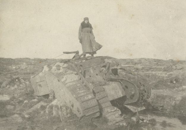 Black and white photograph. A woman stands atop an overturned tank in a rocky landscape. She wears a hat and long coat, which lifts in the wind.