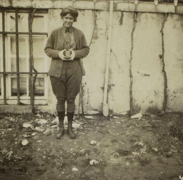 Black and white photograph. A woman smiles at us as she holds a camera in front of her torso. She wears a jacket, button-up shirt, and jodhpur-like trousers over ankle boots.