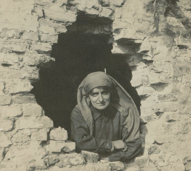 Black and white photograph. A woman leans through a hole in a brick wall. A scarf covers her hair and drapes over her shoulders. She crosses her arms on the opening and looks at us, slightly smiling.