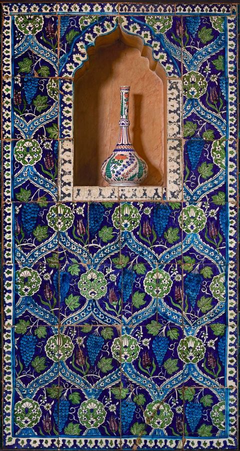 Architecture. Wall tile makes an overarching pattern of continuous S’es surrounding grapes, leaves, and flowers. A blue, white, green, and dark orange vase with a long, narrow neck and flaring body sits in an arched niche near the top.