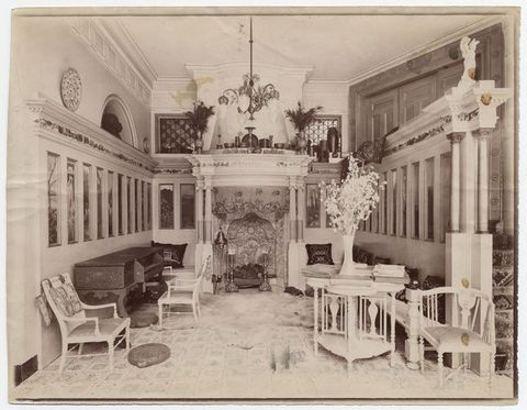 Black and white photograph. A hall-like room is lined with tall, narrow, painted panels in an architectural frame with an embellished entablature. Across from us is an ornate mantlepiece over a richly decorated fireplace. The floor is also patterned, and delicate white furniture is arranged along the edges of the space.
