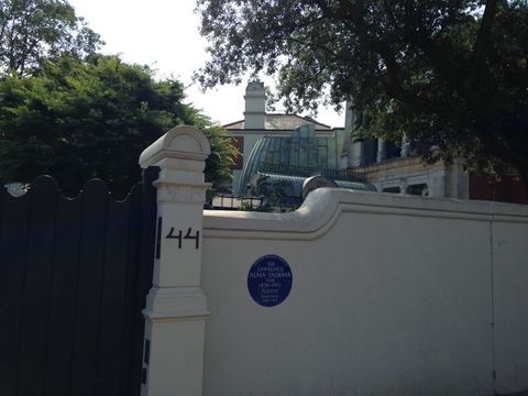 A round blue sign on a white garden in blue text. Trees and rooflines fill the space beyond the wall.