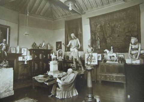 Black and white photograph. A white-haired man sits cross-legged in an armchair in the middle of a room filled with sculpture, paintings, a tapestry, and furniture.