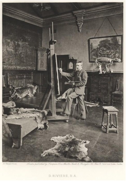 Photogravure. A man wearing a suit stands painting at an easel that nearly reaches the high ceiling. Animal skins drape over furniture and lie on the floor. An animal’s skeleton stands on a piece of furniture on the far wall.