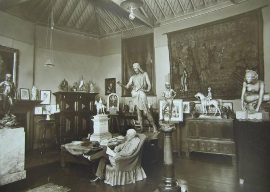 Black and white photograph. A white-haired man sits cross-legged in an armchair in the middle of a room filled with sculpture, paintings, a tapestry, and furniture.