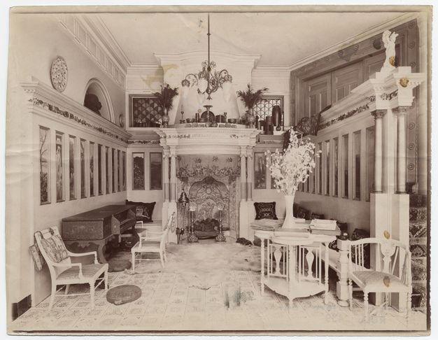 Black and white photograph. A hall-like room is lined with tall, narrow, painted panels in an architectural frame with an embellished entablature. Across from us is an ornate mantlepiece over a richly decorated fireplace. The floor is also patterned, and delicate white furniture is arranged along the edges of the space.