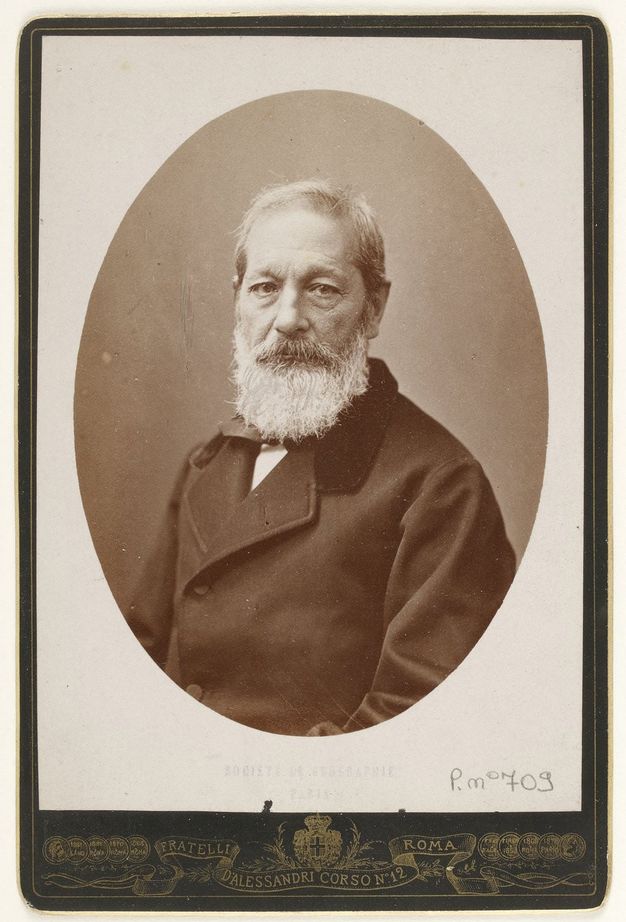 Sepia-toned photograph. In an oval frame, a pale-skinned man with wispy hair and a full white beard looks out at us. His forehead and cheeks are lined with wrinkles.