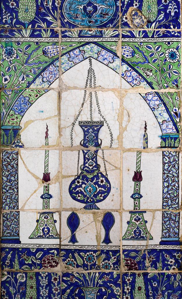 Tiles. Tiled panel shows a round-bellied mosque lamp and two candlesticks under an arch. The objects and patterns on and around the arch are in shades of jewel-toned blue and green.
