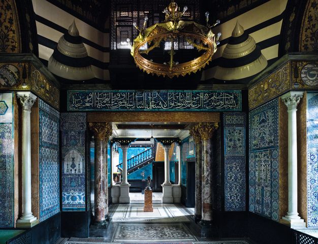 Architecture. Nearly every inch of floor, ceiling, and walls of the space occupy and what is visible along the length of this hall is decorated with intricate, active patterns. The walls are tiled in blue, white, green. A round, gold chandelier hangs from above. The opening leading to the next space is flanked by two pairs of columns with gold capitals. The floors are mosaic.