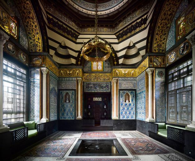 Architecture. Every surface of this room is covered in dense pattern, including the windows. A dome above rises from an octagonal drum, and both overlaid with gold and black designs. Broad arches span windows screened with finely pierced shades. Tiled floors and walls are in tones of blue, red, and gold.
