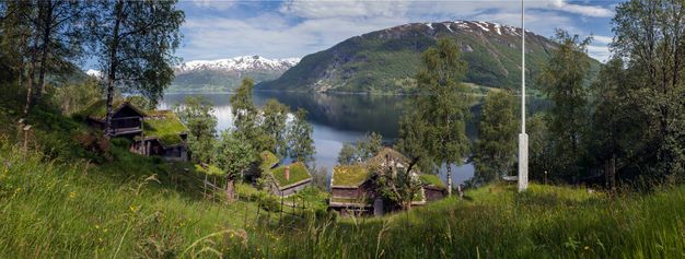 We look down a wildflower-dotted grassy hill at a clutch of buildings with moss-green roofs. The land continues down to the placid surface of a lake. Grass or snow-coated hills rise from the horizon in the near and in the deep distance.