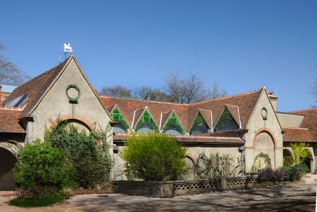 Architecture. This building has a series of gabled windows stretching between two peaked bays. The roof is terracotta brown and the face of the structure tan. In each gable, the space between the arched window below and a triangular peak above is filled with emerald-green tiles.