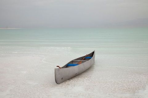 A silver-coloured canoe sits in the shallows of water that deepens from ivory white close to us to pale glacial green in the distance. The horizon is hazy lavender blue.