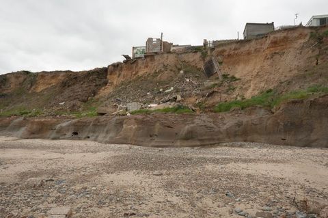 We look down at a coastline alongside steep cliffs. A few buildings are set back from the edge of the cliffs, but a structure or part of one has fallen onto the beach below.