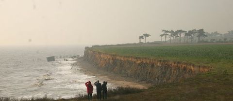 Three people standing in front of us look out at an expanse of steep cliffs that recede away from us. Waves lap at the foot of the cliffs.