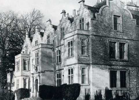 Black and white photograph. A stone manor house is lined with five bracket-shaped gables. Three two-storey projecting bays seem to surge towards sunlight coming from our left.