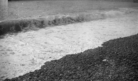 Black and white photograph. A wave breaking onto dark stones makes three distinct, diagonal bands. The lower right corner is the stones. The wide white band of surf has a low swell along the back edge. The water beyond is a midtone grey.