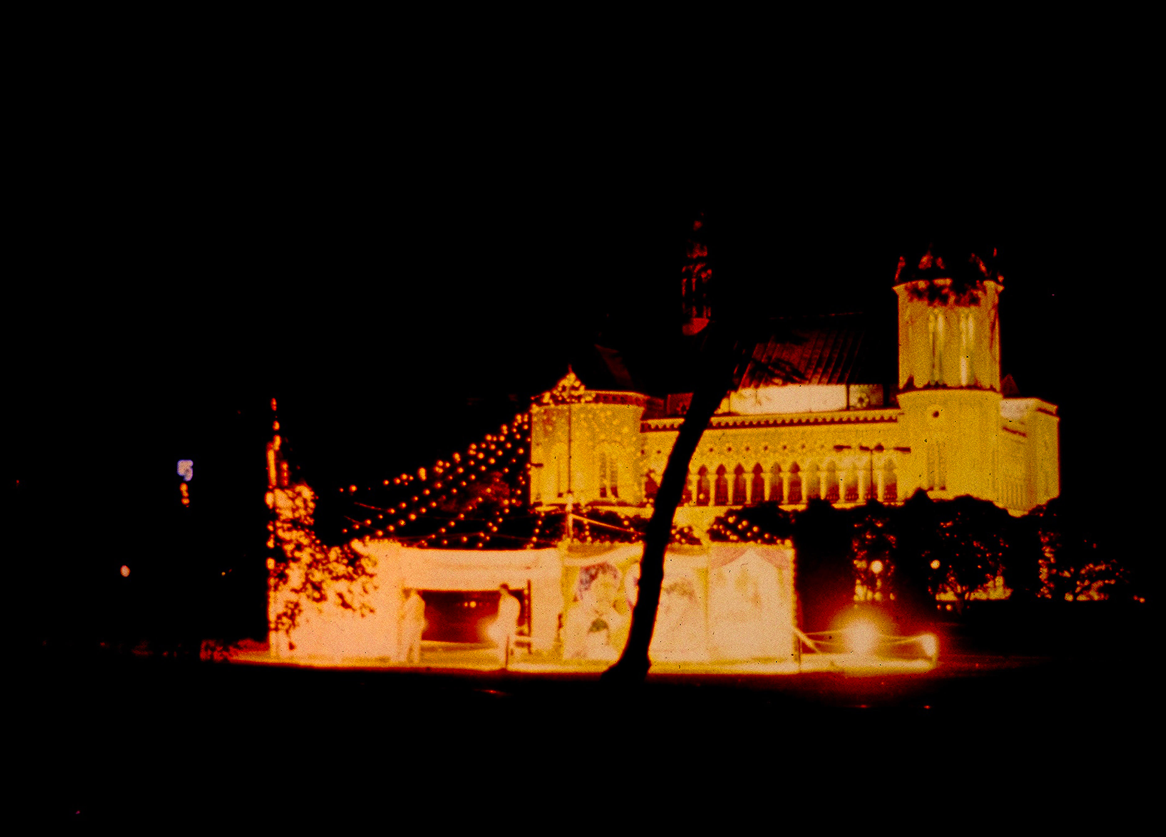 Two structures are lit in amber-yellow light against an ink-black night. A hall with a tower at one end and a row of pointed arches along its length is in the distance. Another structure, like a temporary wall, is closer to us.