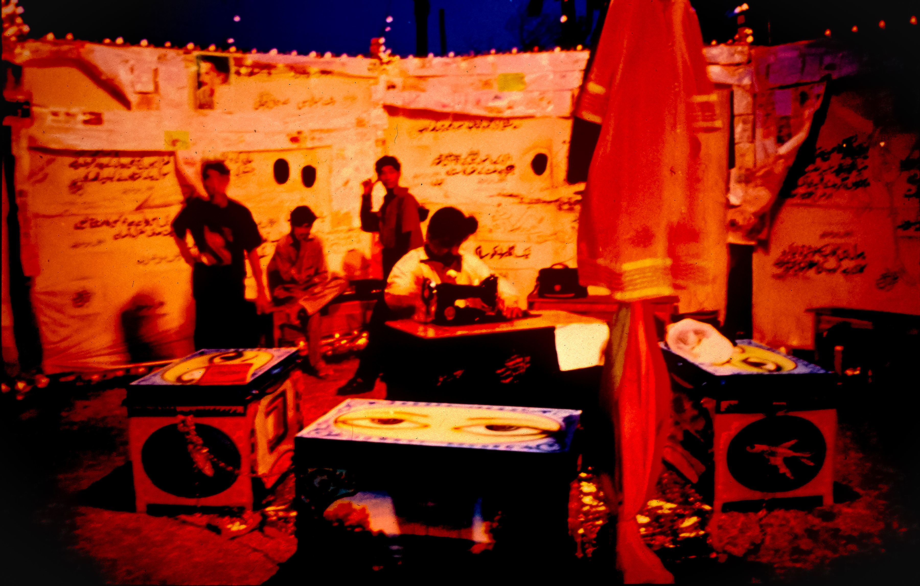 An interior is lit in marigold orange and flame red. A person sits at a sewing machine near the middle of the space. Three more people are behind. The walls appear to have writing and images. The photograph is blurry so details are difficult to make out.