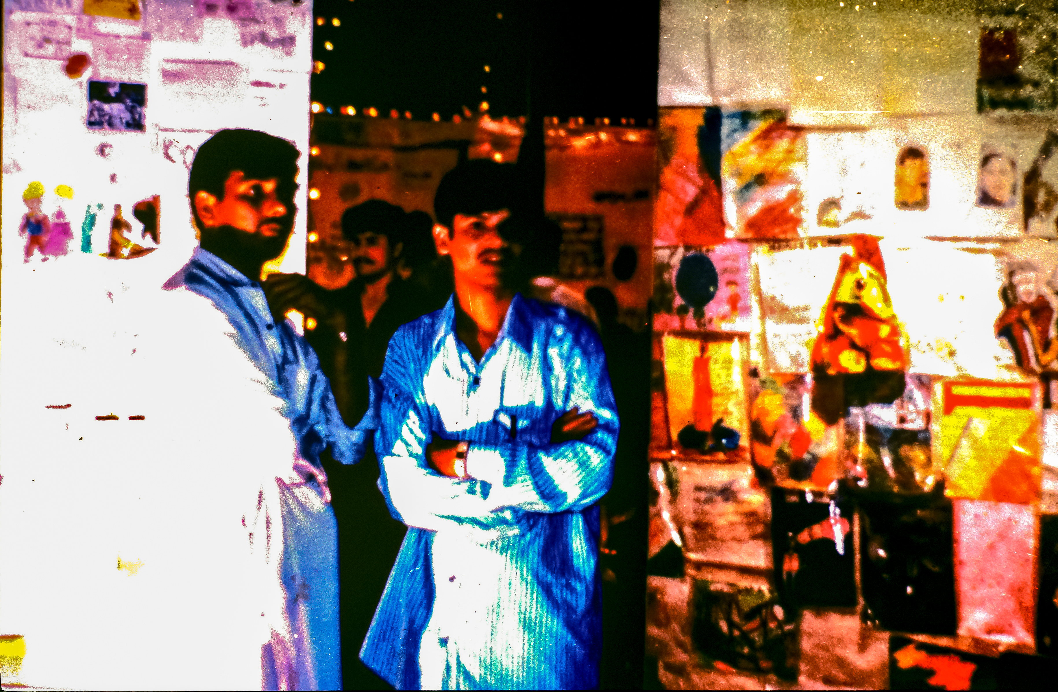 Two men with medium skin, black hair, and moustaches stand in front of walls that appear to be covered with images.
