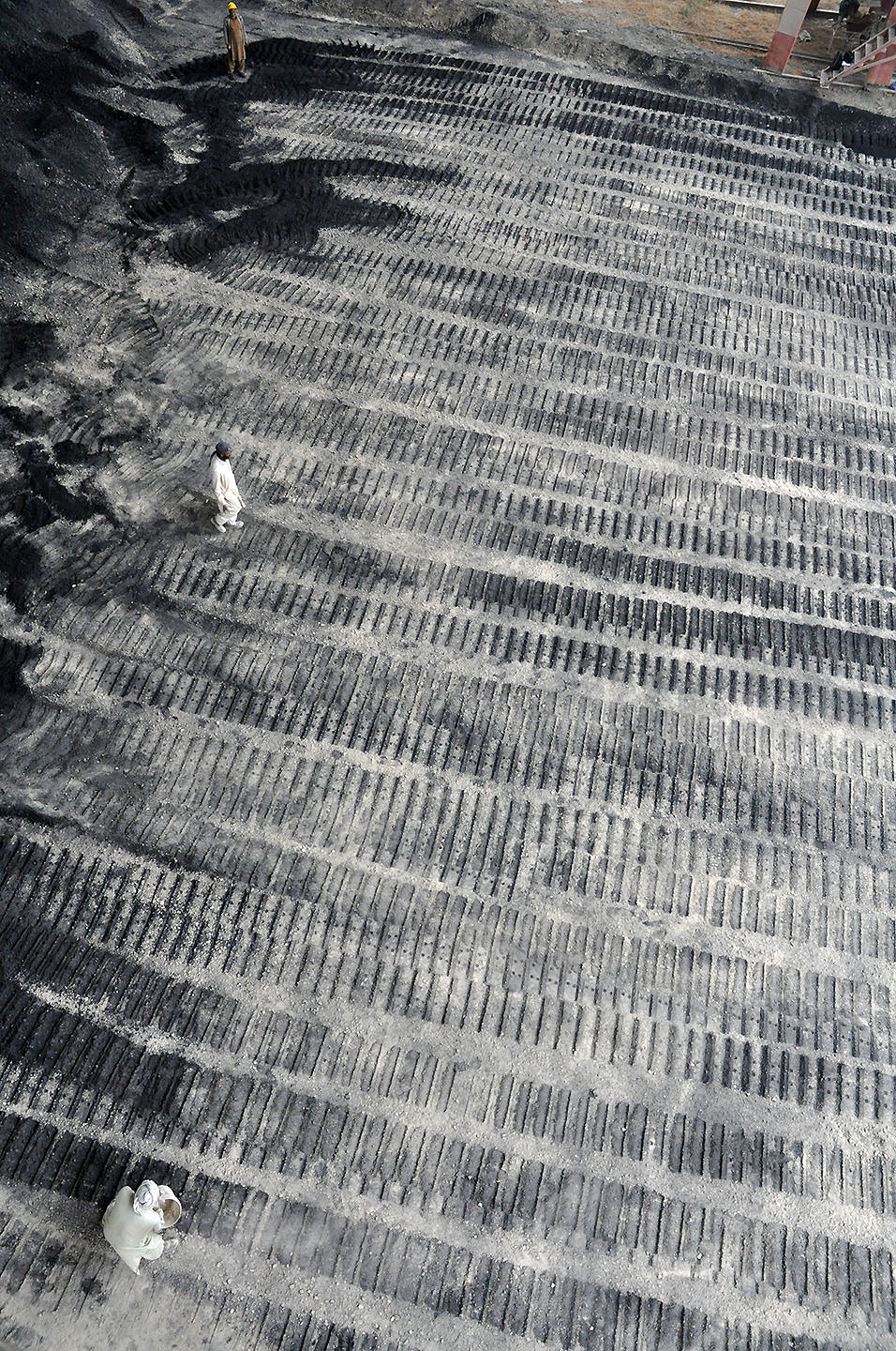 The tracks of a bulldozer’s wheels are imprinted in grey stone dust. We look down on the prepared ground, which resembles rows of zipper teeth. Three people on the ground are miniscule in scale.