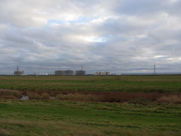 A flat grassland leads to factory buildings and pylons along the horizon.