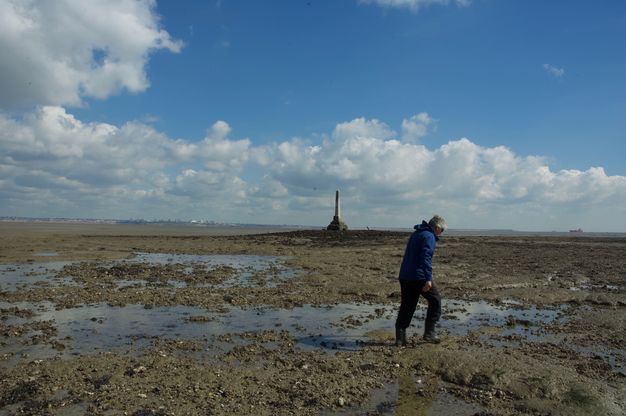 Across a marshy, muddy expanse, an obelisk stands outlined against white clouds in the blue sky at the centre of the image. A person closer to us walks to our right.