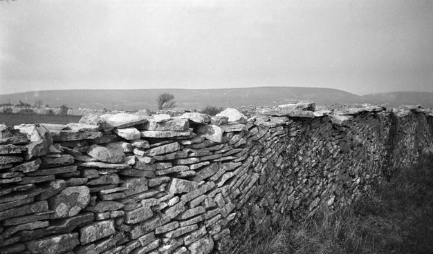 Black and white photograph. Flat rocks stacked to make a wall fall in curves like breaking waves. The wall angles from the lower left corner to the right to cut off our view immediately over the wall. Hills line the horizon in the deep distance.