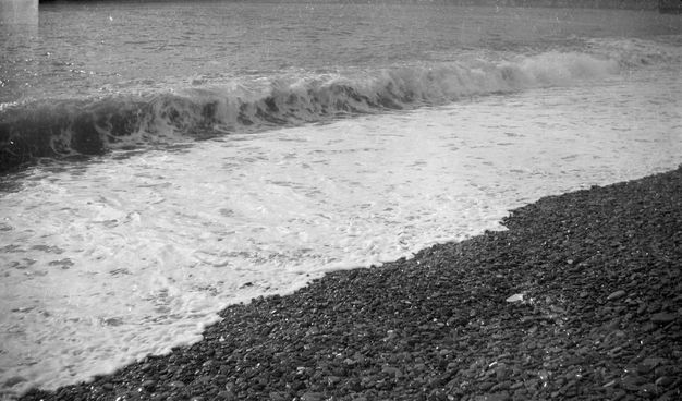 Black and white photograph. A wave breaking onto dark stones makes three distinct, diagonal bands. The lower right corner is the stones. The wide white band of surf has a low swell along the back edge. The water beyond is a midtone grey.