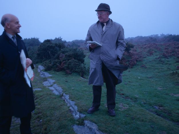Two pale-skinned man stand along a stone line embedded in grass. They wear trench coats and carry notebooks or papers.