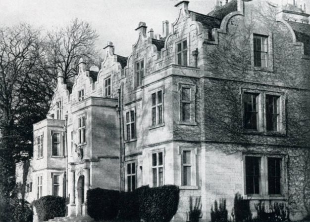 Black and white photograph. A stone manor house is lined with five bracket-shaped gables. Three two-storey projecting bays seem to surge towards sunlight coming from our left.