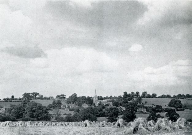 Black and white photograph. A field of haystacks leads to a town of buildings and trees in the lower quarter of this landscape. White clouds march towards us against a pale sky above.