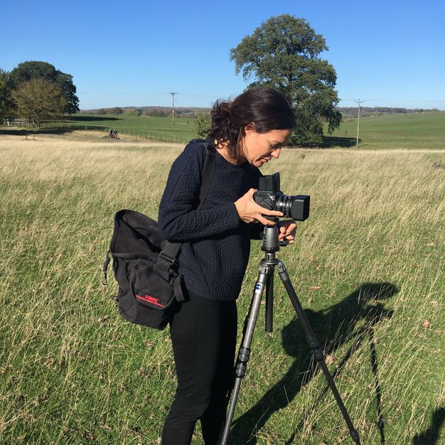 A woman stands behind a camera on a tripod in a grassy field.
