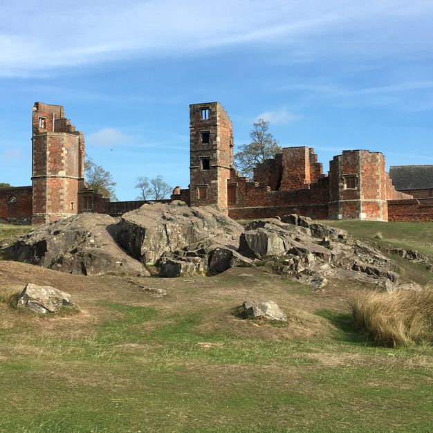 Architecture. The ruins of a red brick building rise beyond a bank of boulders embedded in the field.