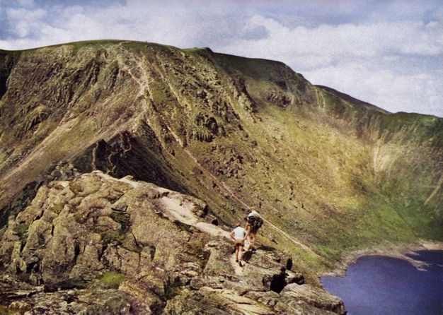 A rocky mountain slides smoothly down to blue water to our right. A short distance from us, two pale-skinned people walk away from us on a path lining a rocky ridge.