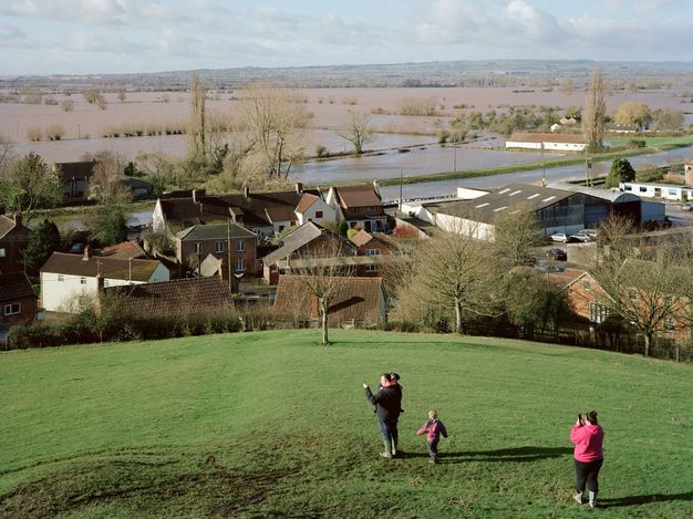 A man, woman, and two children stand on a grassy hill. The adults are looking at their smartphones, presumably taking pictures. A cluster of rooflines at the edge of the hill leads back to an area of flooded fields beyond.