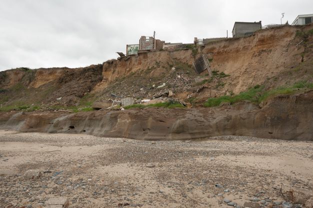 We look down at a coastline alongside steep cliffs. A few buildings are set back from the edge of the cliffs, but a structure or part of one has fallen onto the beach below.