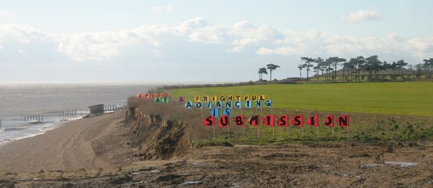 Installation. Steep, low cliffs face a beach to our left. Coloured flags are planted in lines moving back from the edge of the cliff. Each flag has a letter on it. The letters spell out words, and the flags that make up a word have the same colour. Together the flags read, “Submission is advancing at a frightful speed”.
